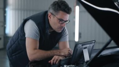 In a car service a mechanic in glasses inspecting and checking the car using a tablet computer that, standing on the opened hood of the car