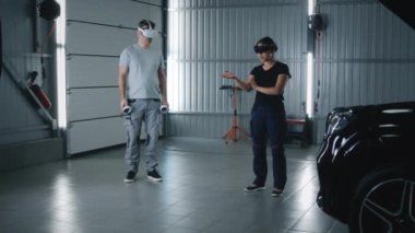 Man mechanic and female asian woman mechanic in virtual reality headsets with controllers talking while checking and diagnosing a car, with a malfunction in a car service