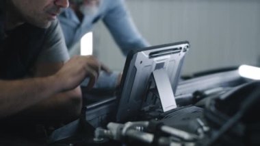 Car service mechanic in repair shop clicking on a screen of digital tablet computer while standing next to a client, near the opened bonnet of the car during diagnostics and maintenance