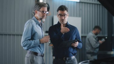 Günlük giysiler içinde araba servisinde bir erkek müşteriyle konuşan ve bir arabayı kontrol edip teşhis koymayı tartışan bir yönetici.