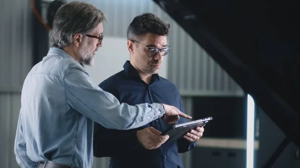 Serious manager in a suit and glasses communicating with a man and diagnosing a car using a gadget in a car service