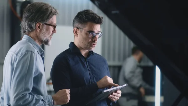 Serious manager in a suit and glasses communicating with a man and diagnosing a car using a gadget in a car service