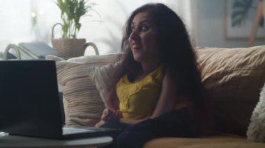 Positive woman with physical disability smiling and communicating online via video call using laptop while sitting on sofa in cozy living room