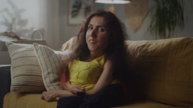 Attractive woman with physical disability smiling and raising her eyebrow while sitting on sofa at home and looking at camera