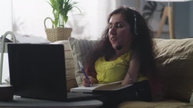 Woman with a physical disability on a sofa at home writing in notebook, while having work video call on a laptop and talking using headset