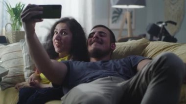 Woman with a physical disability and man laughing and taking a selfie together on smartphone for social networks sitting on a couch at home