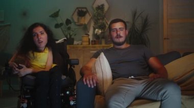 Woman with spinal muscular atrophy sitting in a wheelchair next to her husband and they watching news on TV in the evening together