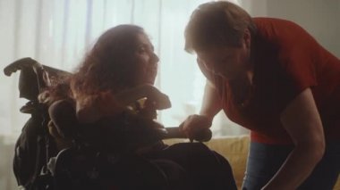Aged woman caregiver helping woman with a spinal muscular atrophy setting a modern motorized wheelchair for comfortable sitting