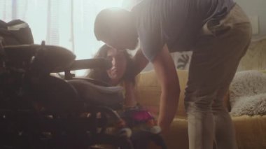 Husband or caregiver taking his wife with spinal muscular atrophy on his hands and helping her to sit in modern motorized wheelchair, together with caregiver