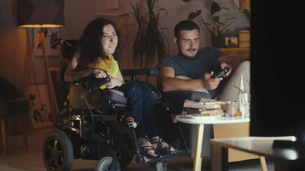 Man playing in video game console on TV using gamepad and woman with a physical disability sitting in a wheelchair at watching it