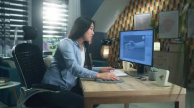 Şaşırmış Asyalı kadın, ofisteki masada oturup bilgisayardaki 3D modelleme projesinde çalışırken kameraya bakıp seviniyor.