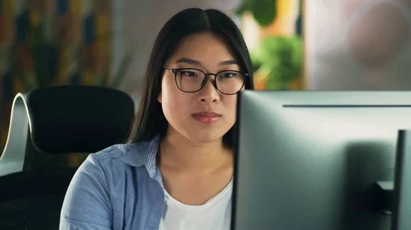 Asian woman in glasses looking at screen of computer while working on 3D modeling project or design remotely from home or surfing the internet in spare time. Freelance