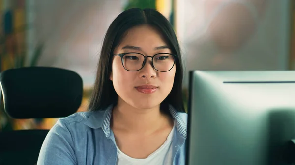Asian woman in glasses looking at screen of computer while working on 3D modeling project or design remotely from home or surfing the internet in spare time. Freelance