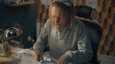 Genç yaratıcı tasarımcı çalışma masasına doğru yürür ev tabanlı bir stüdyoda. Kadın sandalyeye oturur, gözlüklerini takar ve kalemle çizim yapmaya başlar..