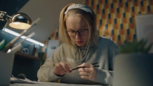 焦点を当てたイラストレーターは彼女のプロジェクトのためにスケッチを描く 灰色のパーカーに身を包んだ眼鏡をかけ 作業机の上に座る若い女性アーティスト 鉛筆のスケッチを作る 物語の概念 — ストック動画
