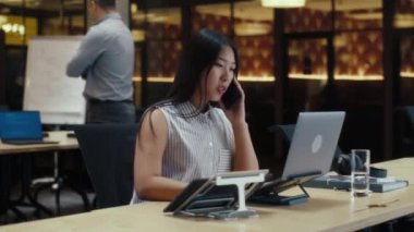 Modern ofiste iş arkadaşlarının yanında çalışırken bilgisayarını ve dijital tabletini kullanan Asyalı bir kadın müşteriyle telefonla konuşuyor. İş kadını e-ticaret girişimini tartışıyor. Şirketin stratejisi.