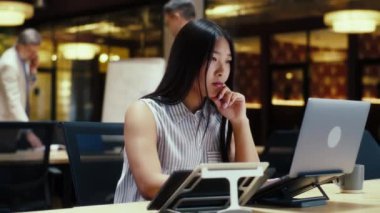 Modern ofisteki Asyalı bir kadın bilgisayar ve akıllı telefon kullanarak iş arkadaşlarının yanında çalışıyor. E-ticaret pazarlama şirketi