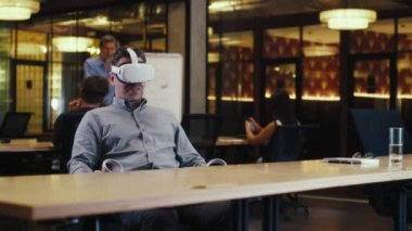 VR kulaklık ve kablosuz kontrolör kullanan, müşteriyle online konuşan, video görüşmesi yapan, yüksek teknoloji şirketinin modern ofisinde çalışan bir adam. İş sanal toplantısı. Siber uzay dijital teknolojisi