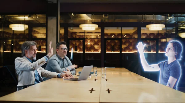 The concept of futuristic business collaborations and negotiations. Two adult businessmen in casual clothes connect and have an online meeting with a young employee using a metaverse holographic link