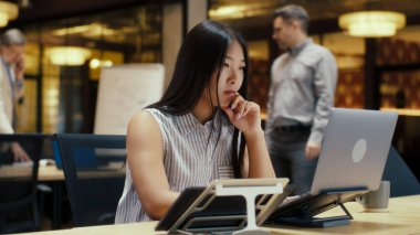 Modern ofisteki Asyalı bir kadın bilgisayar ve akıllı telefon kullanarak iş arkadaşlarının yanında çalışıyor. E-ticaret pazarlama şirketi