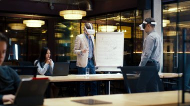 VR kulaklıklarındaki çok ırklı mühendisler, dizüstü bilgisayar ve kablosuz kontrol cihazları kullanıyorlar, planları inceliyorlar, verileri izliyorlar, yüksek teknoloji şirketinin modern ofisinde çalışıyorlar. Siber uzay dijital teknolojisi