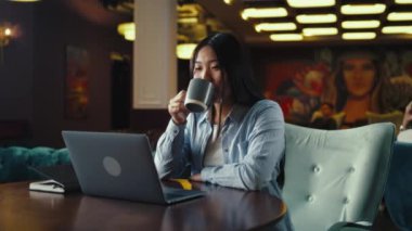 Asyalı iş kadını dizüstü bilgisayar ekranına bakıyor, iş eğitimi izliyor, başlangıç projesinde çalışıyor, ofis kafesinde kahve içiyor. E-ticaret pazarlama şirketi çalışanı.