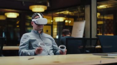 Erkek ofis çalışanı sanal gerçeklikte VR kulaklık ve kablosuz kontrolör kullanıyor, el kol hareketi yapıyor, veri izliyor. Yüksek teknoloji şirketinin modern ofisinde, iş arkadaşlarının yanında çalışmak. Siber uzay dijital teknolojisi