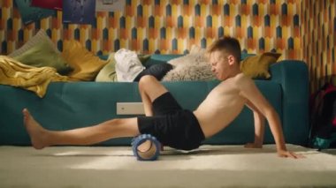 Genç sporcu çocuk odasında köpük silindir kullanıyor. Spor antrenmanlarını başlat. Okul spor takımı futbolcusu. Kasları esnetiyorum. Evde egzersiz yap..