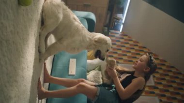 Genç kız yumuşak halıda köpek köpek oyuncağıyla oynuyor, gülümsüyor ve gülüyor, yavru köpekle eğleniyor, evde boş zaman geçiriyor. Dikey çekim. Telefon ekranı görünümü.