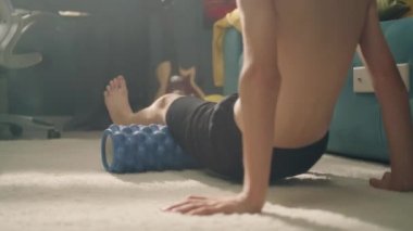 Arka planda, odasındaki halıya kaslarını germek için köpük silindiri kullanan genç bir atlet var. Spor antrenmanlarını başlat. Çıplak vücutlu genç bir çocuk. Okul spor takımı futbolcusu.