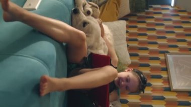 Genç kız kanepede köpekle oturuyor, tablette film izliyor, yavru köpeğe sarılıyor, gülümsüyor ve eğleniyor, evde boş zaman geçiriyor. Dikey çekim. Telefon ekranı görüntüsü. Golden retriever.