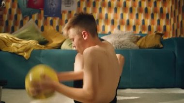 Atletik genç çocuk elinde bir futbol topuyla üst vücut kasları egzersizi yapıyor. Odasında, yerde oturuyor. Yaklaşan bir maç için odaklanmış. Spor eğitimi, sağlıklı yaşam tarzı konsepti..