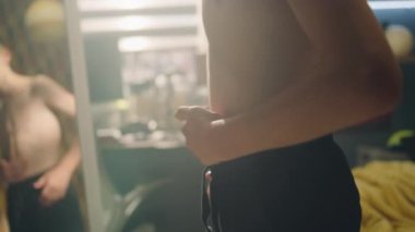 Kararlı genç çocuk odasında egzersiz yaptıktan sonra kaslarını ve kaslarını aynanın önünde inceliyor. Kaslı karın kasları ve omuzları olan çıplak bir vücut. Ağırlık çalışması, fitness, fiziksel aktivite.