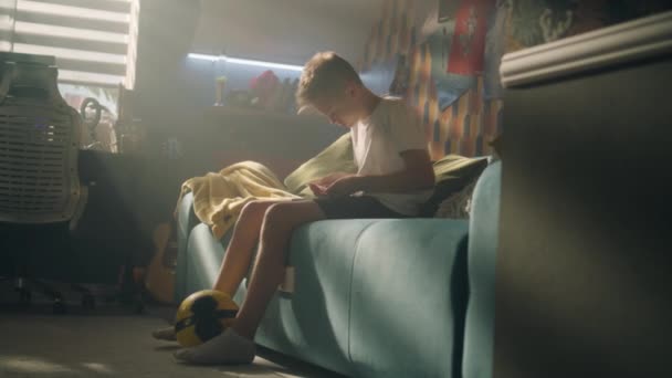 10代の男の子が 打撲した膝に包帯をしている 彼の部屋で一人でベッドに座っている サッカーをしている間に膝を負傷した スポーツ 趣味の概念 — ストック動画