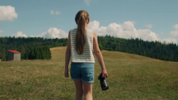 若い写真家が芝生の丘の上にカメラを持って歩く 女の子の美しい風景と素晴らしい自然の景色を見て レジャーの時間を屋外で過ごす スローモーション バックビュー — ストック動画