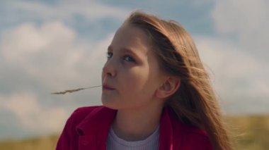 Genç kız oturuyor ve güzel doğayı seyrediyor, ağzında çim tutuyor, düşünceleri serbest bırakıyor ve rahatlıyor, boş zamanlarını dışarıda geçiriyor. Yüzü yakın çekim görüntüsü.
