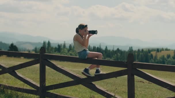 美しい緑の丘の中腹にデジタルカメラを使用して若い写真家の撮影風景は 木製のフェンスに座って 自然の写真を撮る 女の子は素晴らしい写真でポートフォリオを埋める — ストック動画