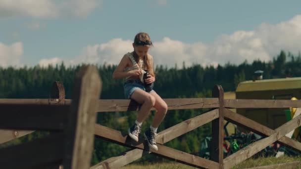 若い写真家のTシャツでデジタルカメラのレンズを拭く 木製のフェンスに座っている 少女は プロセスに集中し レジャー時間を屋外で過ごす美しい自然景観撮影 スローモーション — ストック動画