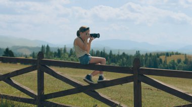Genç fotoğrafçı, güzel yeşil tepe üzerinde dijital kamera kullanarak manzara çekiyor, ahşap çitte oturuyor, doğanın resimlerini çekiyor. Portföyü inanılmaz fotoğraflarla dolduran kız..