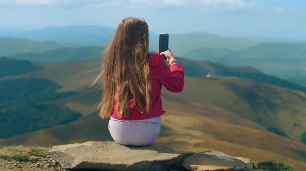 年轻姑娘在美丽的群山之间的山谷里用电话拍摄风景 坐在石头上 在大自然中做出创造性的满足 在户外消磨时光 背景色 — 图库照片