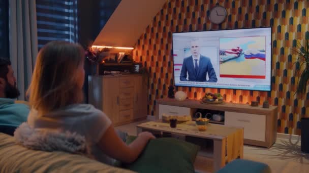 夫妻二人坐在客厅的沙发上吃零食 看电视上有关俄美对抗的新闻 男人和女人晚上呆在家里 电视直播 — 图库视频影像