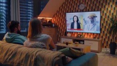 Oturma odasında birlikte oturan çift, modern iç mekanla birlikte televizyon kanalında Covid 19 salgınının yeni dalgasını izliyorlar. Erkek ve kadın hafta sonunu televizyon tıp programında geçiriyorlar.
