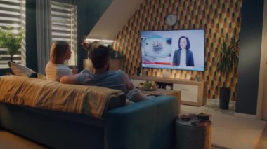 Oturma odasındaki kanepede oturan bir çift çocuk felci ve su kirliliği hakkında TV haberleri izliyor. Erkek ve kadın hafta sonları evde birlikte vakit geçiriyorlar. TV canlı yayın.