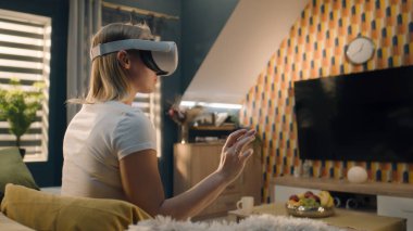 VR kulaklık takan bir kadın, öğle vakti rahat bir oturma odasında kanepede oturuyor, el kol hareketi yapıyor, sanal gerçeklikte video oyunu oynuyor, VR gözlüklerini test ediyor, hafta sonunu evde geçiriyor. Siber uzay ve metaevren.