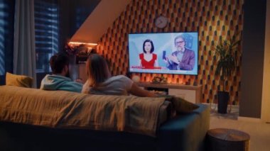 Oturma odasındaki kanepede oturan ve abur cuburlarla televizyon karşısında yeni akıllı telefon sergisini seyreden, hafta sonunu rahat bir evde geçiren çift. TV canlı yayın.