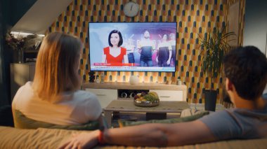 Çift kanepede oturuyor, konuşuyor, canlı yayında siber spor, ünlü siber takım ve oyun turnuvası hakkında haberler izliyor. İşten sonra kadın ve erkek evde dinleniyor. TV canlı yayın.