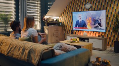 Oturma odasında birlikte oturan çift Ukrayna 'daki savaş ve Rus işgali hakkında TV haberleri izliyor. Erkek ve kadın işten sonra evde vakit geçiriyorlar. TV canlı yayın.