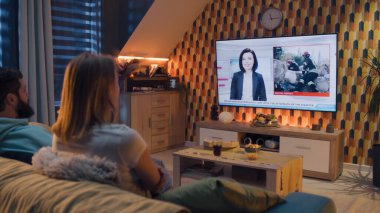 Oturma odasındaki kanepede oturan bir çift. Yanında abur cubur ve gazoz var. TV 'de felaket, terörist saldırı, bina çökmesi ve enkazdan kurtarma haberleri izliyorlar. Evde vakit geçirmek. TV canlı yayında.