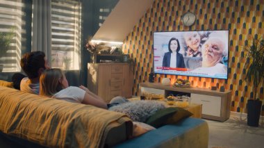 Oturma odasındaki kanepede oturan çift, modern iç mekanla konuşuyor, TV haber programını izliyor, tıptan ve abur cubur ve gazozlu hastalıklardan bahsediyor. Spouces evde birlikte vakit geçiriyor. TV canlı yayın.