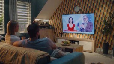 Oturma odasındaki kanepede oturan çift yeni akıllı telefon ya da dijital ürün sunumu hakkında TV haberleri izliyor. Erkek ve kadın evde birlikte vakit geçiriyorlar. TV canlı yayın.